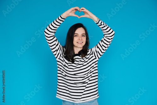 Happy Woman in Striped Sweater Spreading Love and Positivity - Smiling Girl with Raised Arms Forming Heart Shape, Expressing Joy and Affection - Isolated Blue Background - Ideal for Happiness, Love © Yauhen