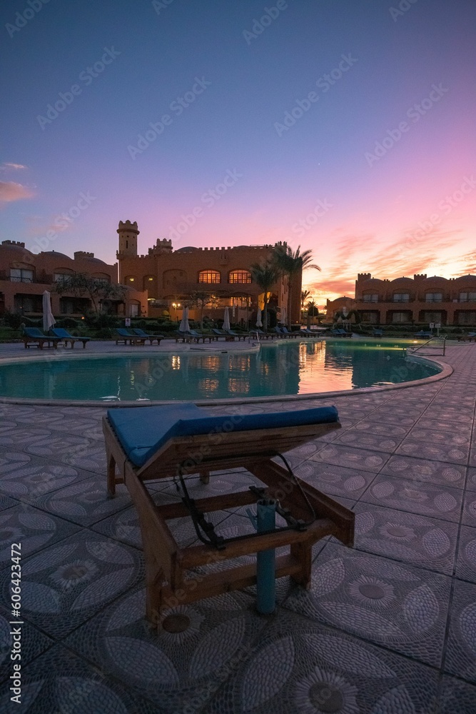 Fototapeta premium Vertical of a blue poolside lounge chair next to a big swimming pool captured at sunset