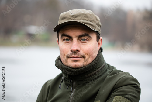 Portrait of smiling military man in olive uniform and cap against lake in rural area on cold winter day © Olena Shvets