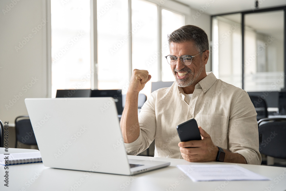 Middle-aged Latin or indian businessman screaming proud cheering with excitement , man read great news on smartphone celebrating victory and success sitting front of laptop computer working at office. Middle-aged Latin or indian businessman screaming proud cheering with excitement , man read great news on smartphone celebrating victory and success sitting front of laptop computer working at office.