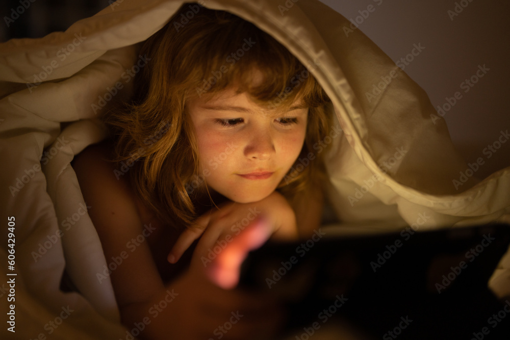 Child gadget addiction and insomnia. Kids with social media. Child lying in bed playing a tablet in dark room, light under blacket. Close up of kid watching cartoons on tablet. Child gadget addiction and insomnia. Kids with social media. Child lying in bed playing a tablet in dark room, light under blacket. Close up of kid watching cartoons on tablet.