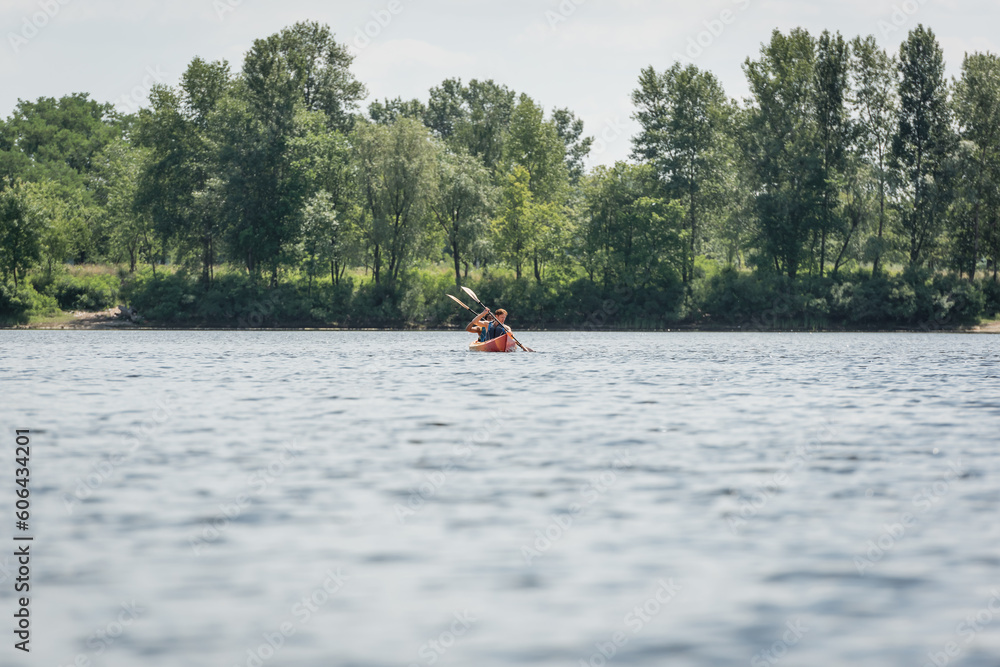 view from afar of active and interracial couple in life vests sailing in sportive kayak during recreation weekend on river with green bank during water recreation in summer