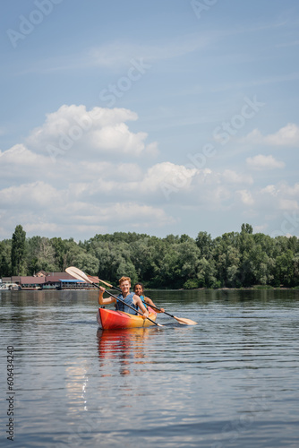 sportive multiethnic couple in life vests paddling in kayak on river with green picturesque bank under blue cloudy sky during recreation weekend in summer