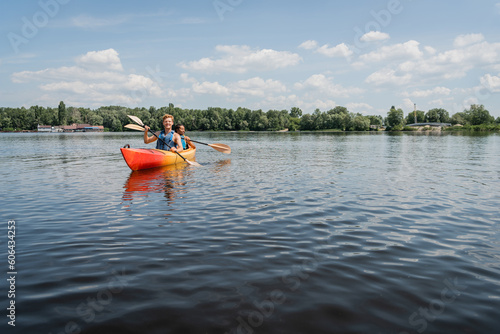 active multiethnic couple in safe vests sailing in sportive kayak on picturesque lake with green shore under blue and cloudy sky during water recreation on summer weekend © LIGHTFIELD STUDIOS