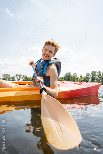 cheerful and redhead man in life vest holding paddle and looking at camera while spending time on lake by sailing in kayak during water recreation on summer weekend