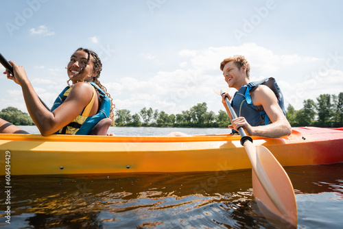 smiling and pretty african american woman sailing in sportive kayak with active redhead man in life vest during summer vacation on river with green bank