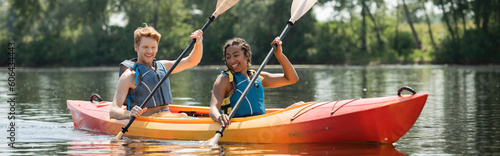 smiling and active multiethnic friends in life vests paddling in sportive kayak during water recreation weekend on picturesque river on summer day, banner photo