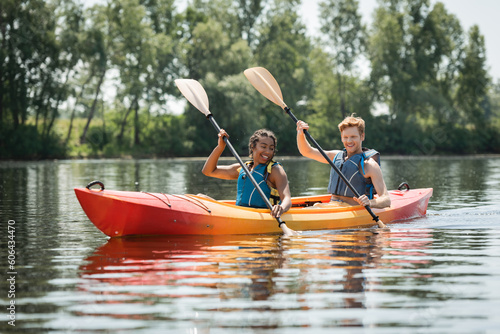 charming african american woman with young and redhead man in life vests smiling while paddling in sportive kayak on lake with green trees on shore in summer