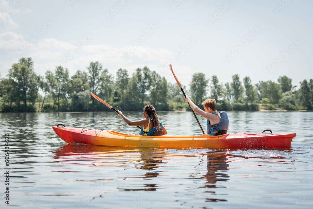 side view of active multiethnic couple in life vests holding paddles while sailing in sportive kayak on river with picturesque bank with green trees in summer side view of active multiethnic couple in life vests holding paddles while sailing in sportive kayak on river with picturesque bank with green trees in summer
