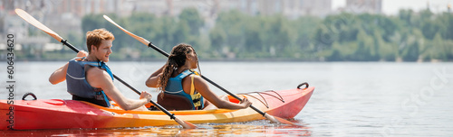 young and redhead man and african american woman in life vests sailing in sportive kayak with paddles on calm river during water recreation on summer day, banner