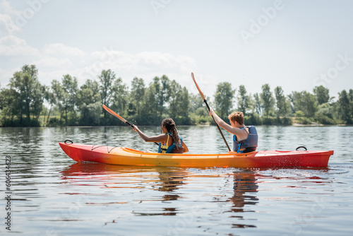 side view of active multiethnic couple in life vests holding paddles while sailing in sportive kayak on river with picturesque bank with green trees in summer