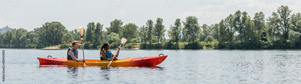 side view of young man and african american woman in life vests sailing in sportive kayak during active summer weekend on picturesque river with green trees on bank, banner
