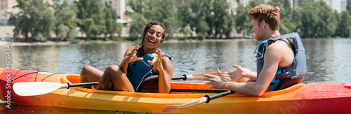 cheerful african american woman in life vest gesturing near young redhead man while spending summer weekend on river and sitting in sportive kayak, banner
