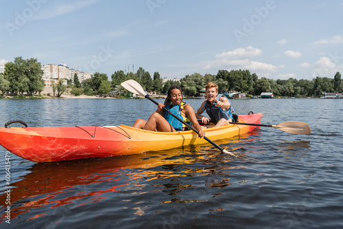 young and happy redhead man in life vest pointing with finger and paddling in kayak with enchanting african american woman on lake with green shore in summer