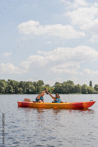 carefree interracial couple in life vests giving high five while spending summer weekend on river and sailing in sportive kayak along green riverside under blue sky with white clouds © LIGHTFIELD STUDIOS
