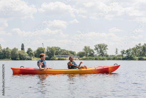 side view of joyful african american woman and young redhead man in life vests sailing in sportive kayak on river with green bank under blue and cloudy sky © LIGHTFIELD STUDIOS