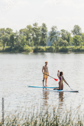 redhead and sportive man paddleboarding along green riverside near african american woman in colorful swimsuit during summer recreation on summer weekend © LIGHTFIELD STUDIOS