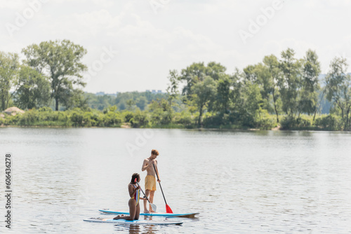 active multiethnic couple in colorful swimwear sailing on sup boards with paddles while spending summer vacation day on river with green trees on scenic bank