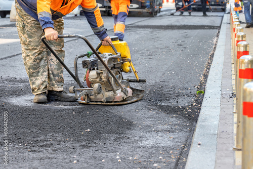 A road service worker compacts fresh asphalt with a gasoline vibrating plate around a sewer manhole on the road.
