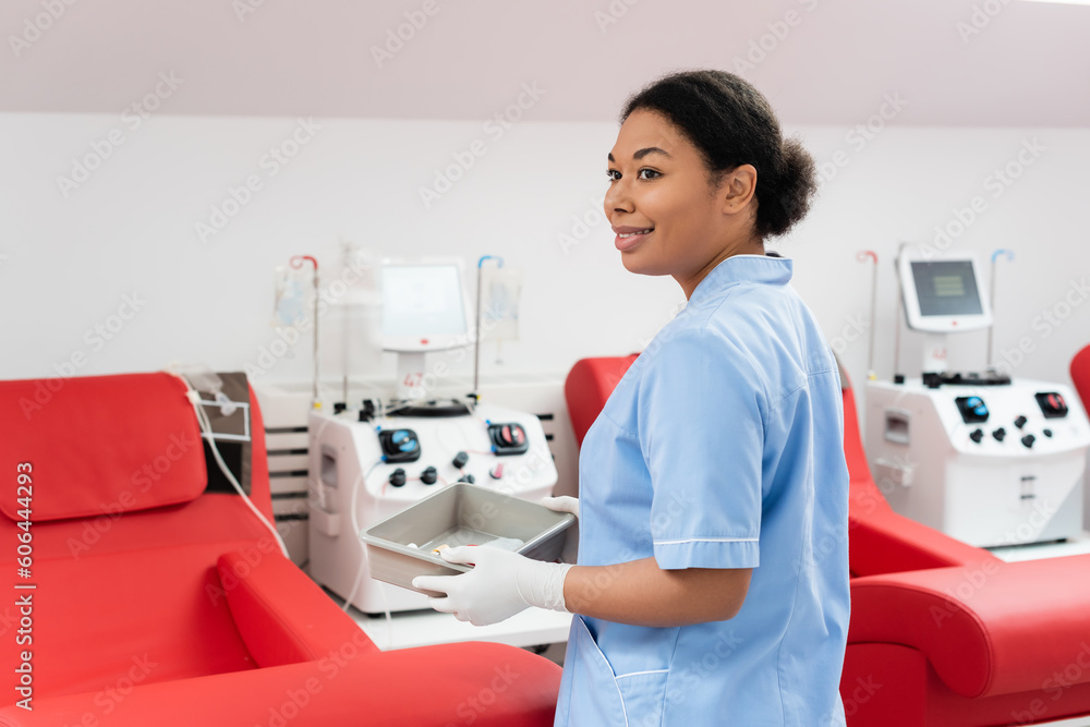 Obraz premium smiling multiracial nurse in blue uniform and latex gloves holding medical tray near transfusion machines and ergonomic chairs in blood donation center
