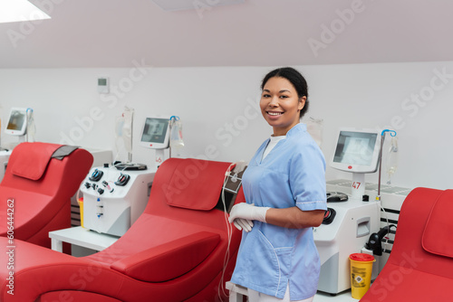 joyful multiracial nurse in blue uniform and latex gloves looking at camera near medical chairs and transfusion machines with monitors in blood donation center