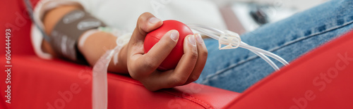 partial view of multiracial woman with transfusion set holding rubber ball while sitting on ergonomic medical chair during blood donation in laboratory, banner