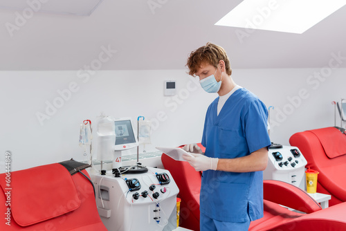 redhead doctor in blue uniform  medical mask and latex gloves looking at digital tablet near medical chairs and automated transfusion machines in blood donation center