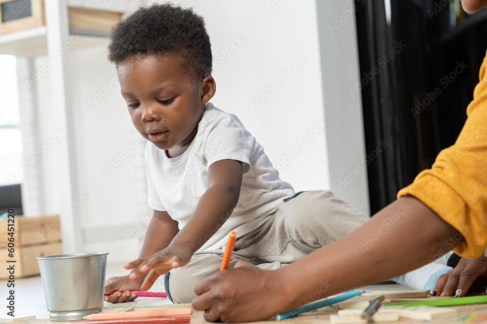 Φωτογραφία Cheerful African-American mother and cute toddler son playing with colored pencils, drawing on a big piece of paper, and sitting on the floor