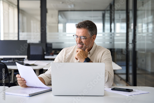 Focused Hispanic businessman doing paperwork in office with copy space. Happy Latin or Indian male business man holding documents, working at laptop computer doing online trade market tech research.