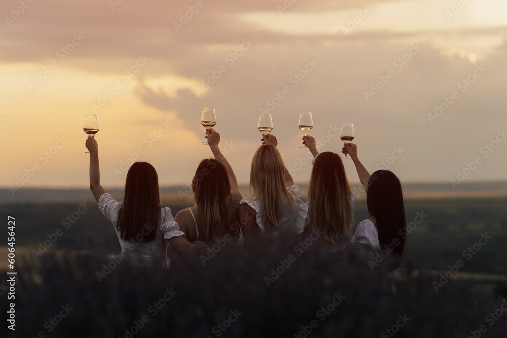 Back view of company of young female friends with hands up having fun, raising glasses with wine and enjoy beautiful sunset at summer picnic in lavender field.