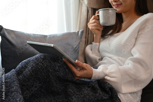 Cropped image of Young Asian woman using tablet and holding a cup of coffee while sitting at the couch with blanket.