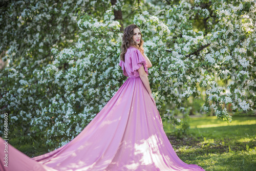 A young beautiful brunette girl stands near a branchy spring apple tree. A girl in a pink dress smiles between blooming apple trees. The concept of spring or summer