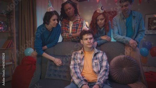 A curlyhaired guy, surrounded by his friends in party hats, sits upset on the couch because of the defeat in the game. Friends support him and encourage him. Games, entertainment at home parties