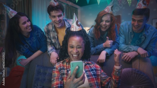 An African American woman is playing an online game on her phone while her friends are watching her from behind. The girl wins, waving her hand in a victory gesture, friends show a thumbs up, class.