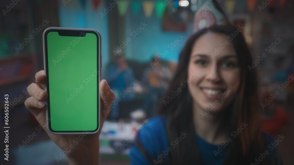 A girl in a festive hat shows a smartphone with a green screen, smiles. Close up of green screen smartphone on a blurred background of a party, in a room decorated with a disco ball, flags.