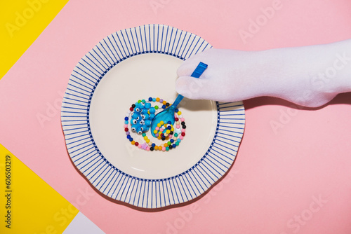 A foot in a white tabi sock holds a blue plastic spoon over colored necklace beads in the form of food over a large plate. Creative concept, top view