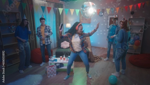 A group of friends are dancing in a room decorated with a party. An African American girl in red wireless headphones dances to her playlist on her phone. Party, birthday, celebration.
