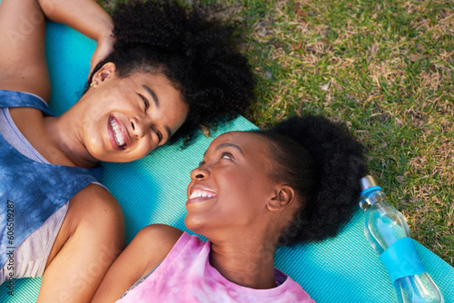 Two young Black women lie on yoga mat after exercise in park, relaxing talking