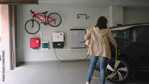 The woman parks in the garage, plugs a charger into the electric car socket. Green energy from electric vehicles for healthy environment. Eco power from renewable source at home. Horizontal 4k video.