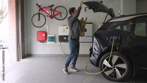 Man multitasking arrives home after shopping at the grocery store and plugs a charger to electric car socket at garage. Renewable houses, Energy Efficient Domestic Life. Horizontal Video 4k.