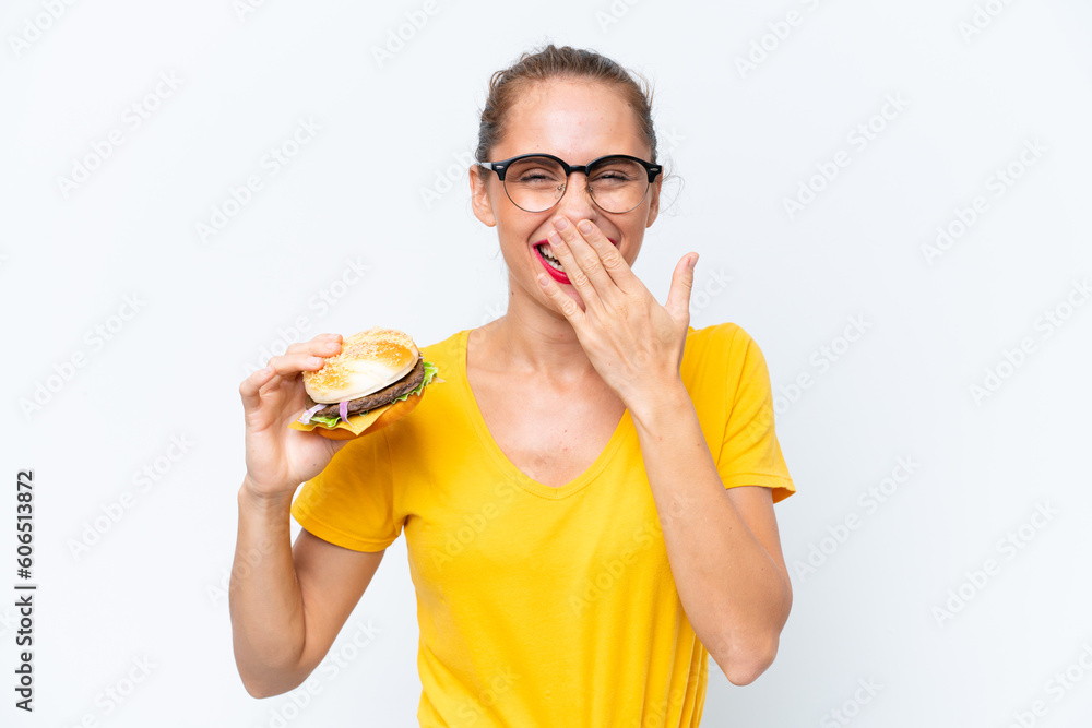 Young caucasian woman holding a burger isolated on white background happy and smiling covering mouth with hand Young caucasian woman holding a burger isolated on white background happy and smiling covering mouth with hand