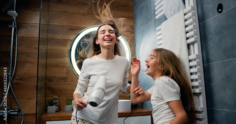 Funny young woman and 10 years old girl with long hair in pajamas drying her voluminous hair by hair dryer while dancing laughing and looking at each other in modern bathroom in home - obrazy, fototapety, plakaty 