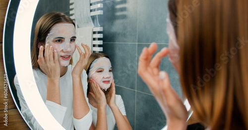 Portrait of two beautiful girls with facial face masks cosmetic looking at the mirror while standing in the bathroom at home. Skin care and beauty concept