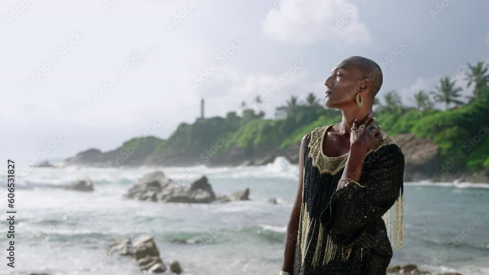 Vivid queer black male in luxury gown poses on scenic ocean beach. Trans sexual ethnic fashion model in long posh dress and accessories looks at camera, touches neck with hand slow motion Vivid queer black male in luxury gown poses on scenic ocean beach. Trans sexual ethnic fashion model in long posh dress and accessories looks at camera, touches neck with hand slow motion