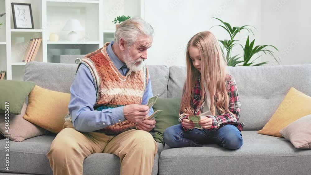 Generous aged gentleman sitting on grey couch and giving some cash to petite golden-haired girl for excellent job around house. Concept of relationships between grandfather and granddaughter. Generous aged gentleman sitting on grey couch and giving some cash to petite golden-haired girl for excellent job around house. Concept of relationships between grandfather and granddaughter.