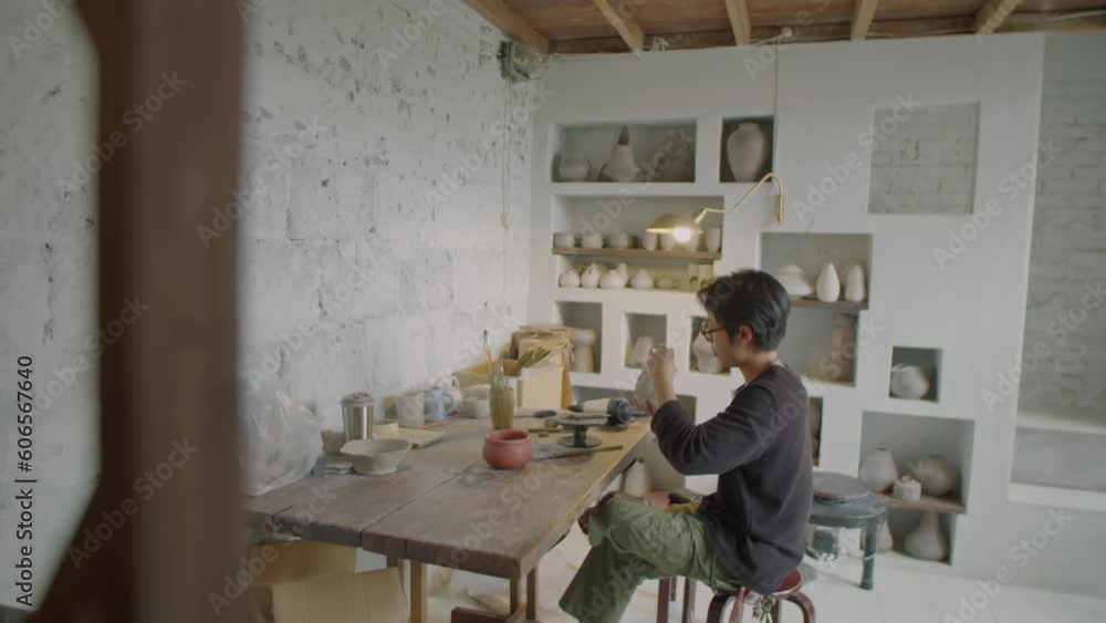 Full shot of young male artisan sitting at workplace in pottery studio and making ceramic mug Full shot of young male artisan sitting at workplace in pottery studio and making ceramic mug