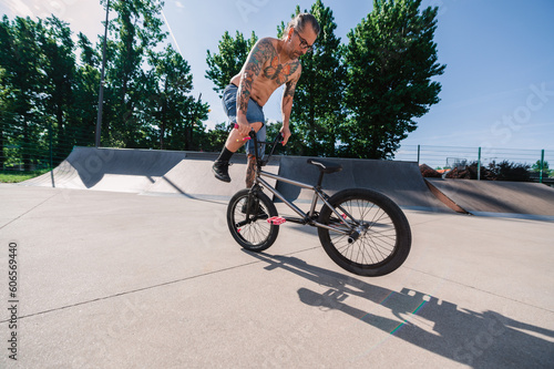 A tattooed shirtless urban guy is performing freestyle tricks and stunts in a skate park.