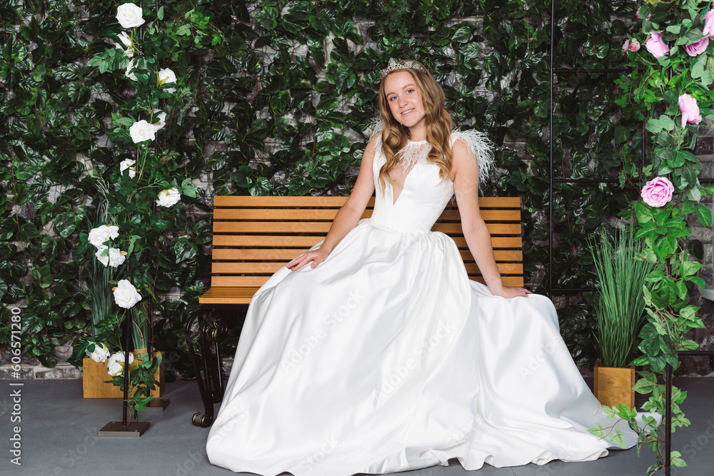 Beautiful bride in elegant white dress and tiara sits on a wooden bench in front of plants and an arch with flowers. Bride in a garden.