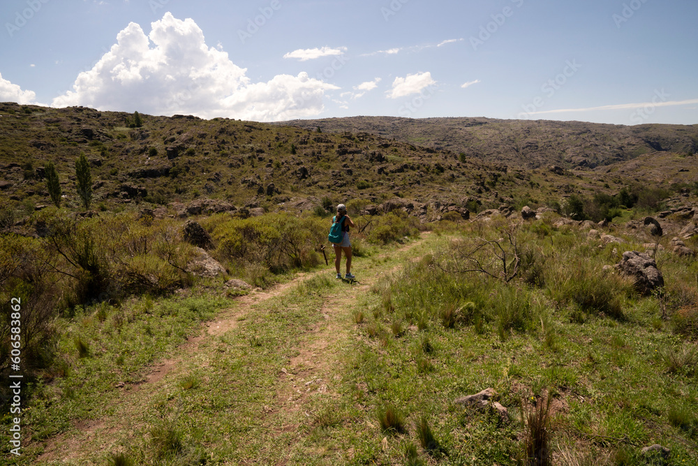 Outdoor activities in a sunny day. View of a young woman walking along the hiking path across the hills and green grassland. 