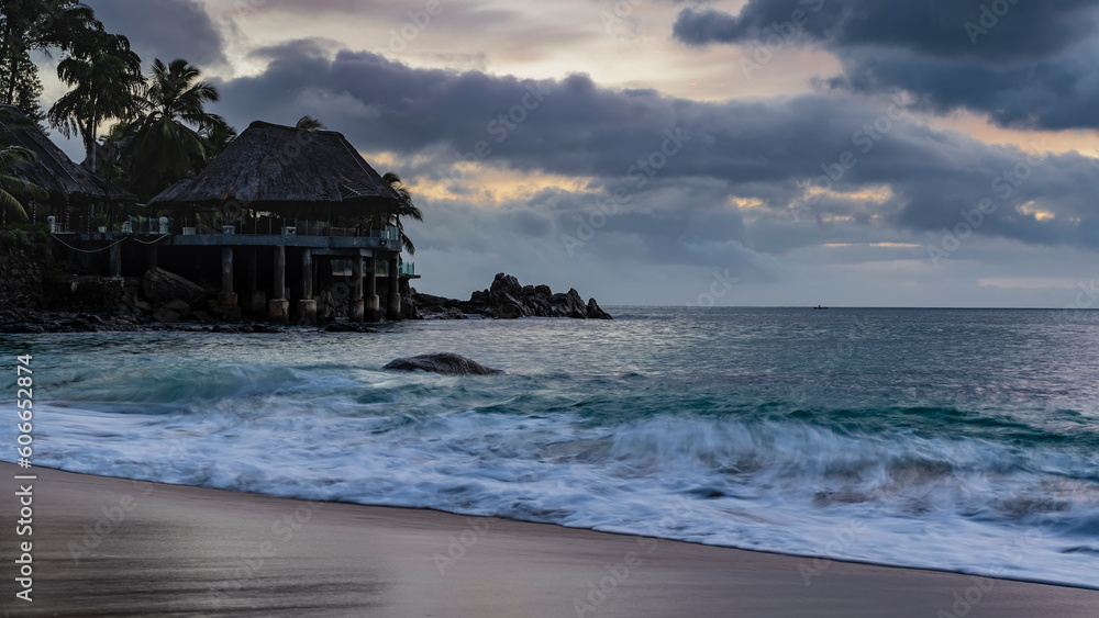 The waves of the turquoise ocean are foaming on the sandy beach. Boulders at the water's edge. Silhouette of a gazebo with a thatched roof against the evening sky, clouds. Long exposure. Seychelles